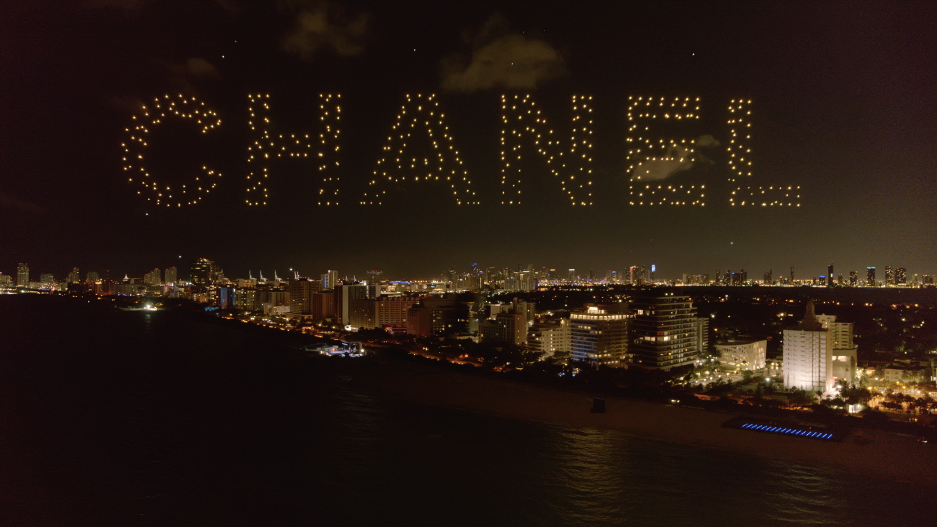 A drone display in the night sky spelling 'Chanel' over a brightly lit city, creating a striking and elegant visual tribute to the iconic brand.