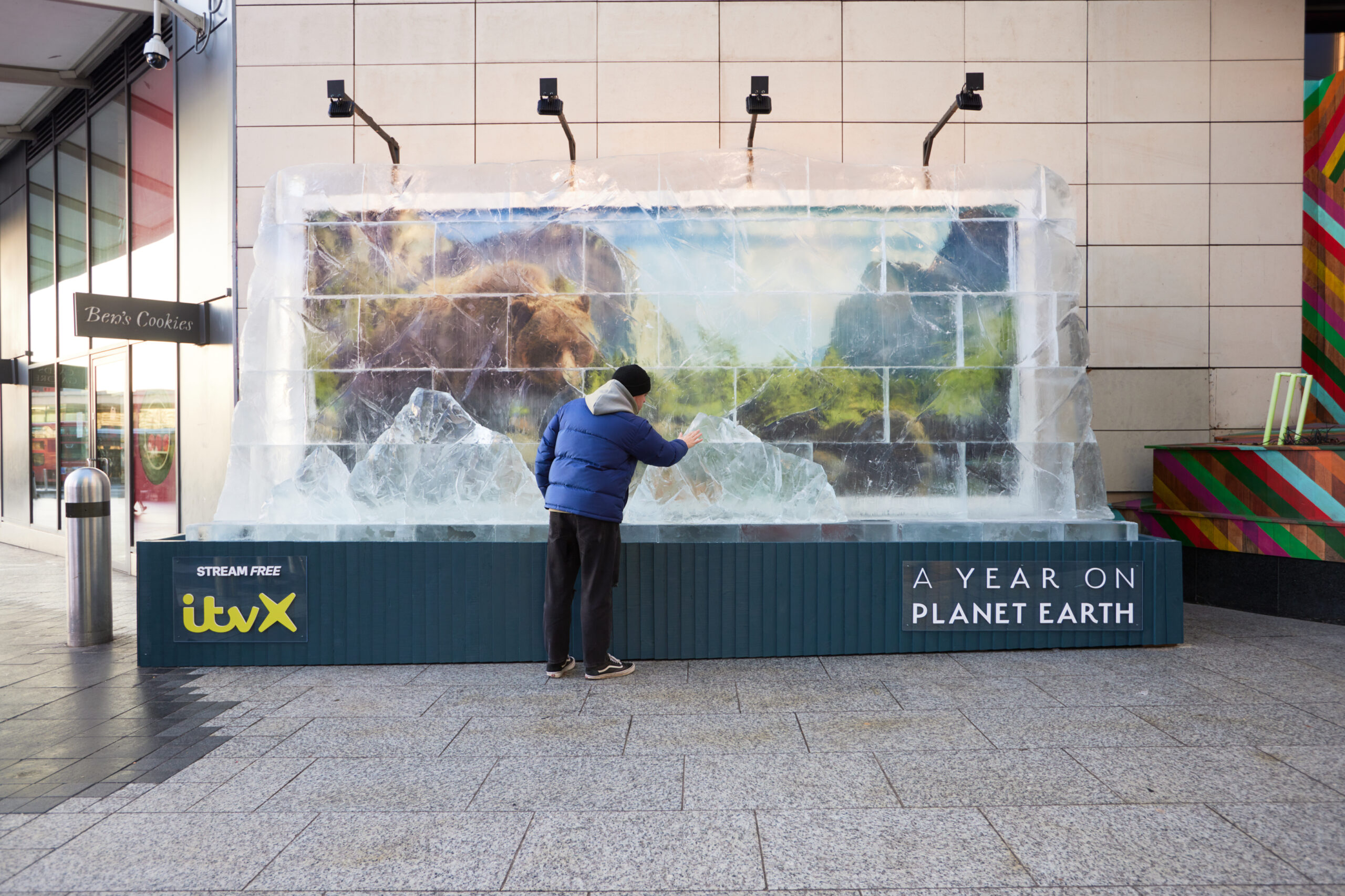 A man in a blue jacket touches a large ice block encasing a billboard for ITVX's 'A Year on Planet Earth' at Westfield London, with the show's title visible at the bottom.