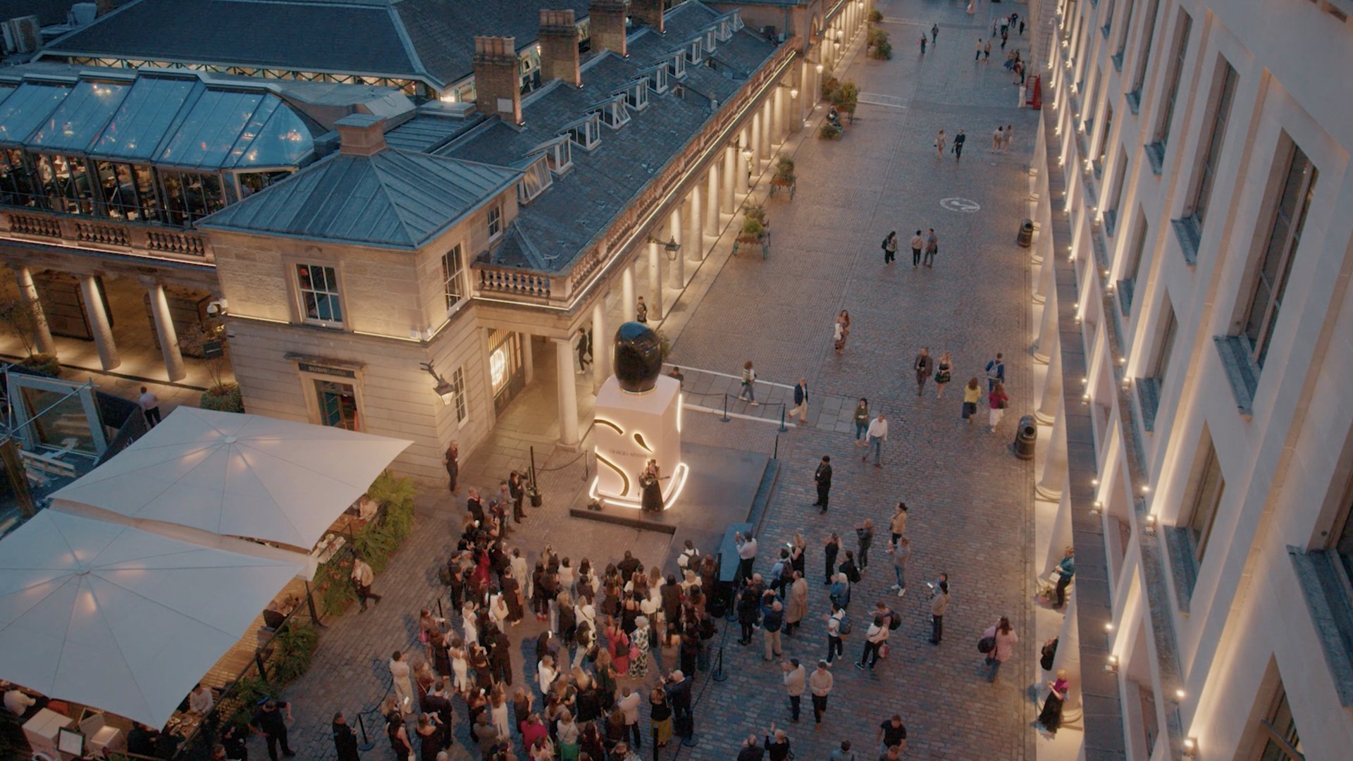 Overhead view of a large crowd gathered around an illuminated Giorgio Armani Sì installation at dusk. A singer performs on stage in front of the crowd, with many attendees holding up phones to record the moment. The installation glows in pink and red tones, creating a striking contrast against the dimming sky and cobblestone surroundings.