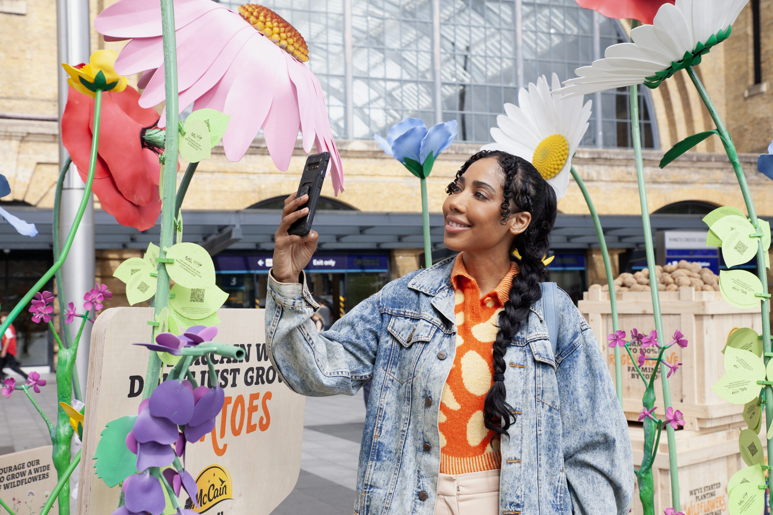 A woman interacting with McCain’s wildflower installation at King’s Cross Station, London. She takes a photo of a large, colourful wildflower model which is part of McCain’s “Let’s All Chip In” campaign promoting regenerative agriculture and sustainability.