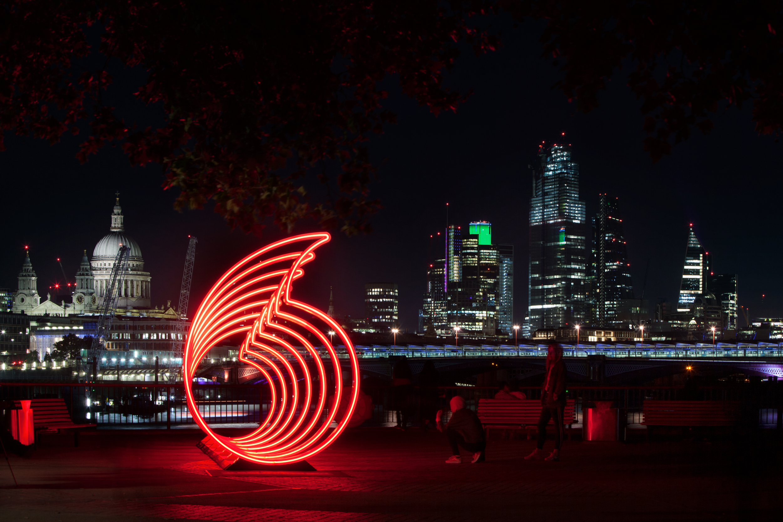 Illuminated Vodafone installation on London's Southbank. 7 life-sized illuminated Vodafone logos are aligned one behind the other, creating the impression of an infinite motif. The image is taken at night, highlighting the installation against the pitch-black sky. In the distance, London's skyline is visible, with buildings lit up and windows glowing.