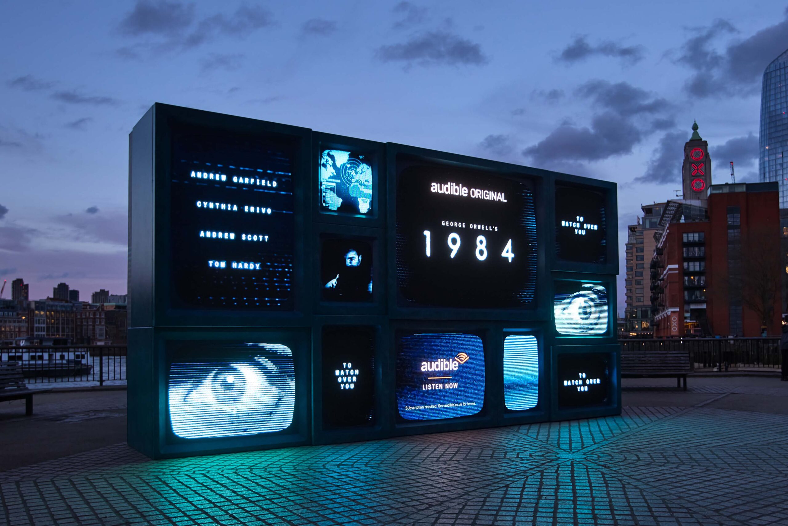 A large blue multi-screen billboard display for Audible's '1984' adaptation in Southbank, London. The billboard features multiple screens resembling old TVs, with text showing names like 'Andrew Garfield' and 'Cynthia Erivo' as well as 'George Orwell’s 1984'. The setup has a gritty, retro design with close-up images of eyes and phrases like 'Every room has one' displayed across various screens.