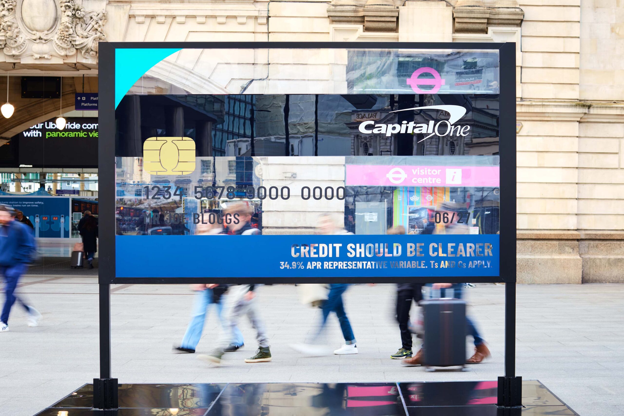 A transparent billboard installation at Victoria Station in London shaped like a large credit card, with people walking by, part of Capital One's 'Credit Made Clearer' campaign.