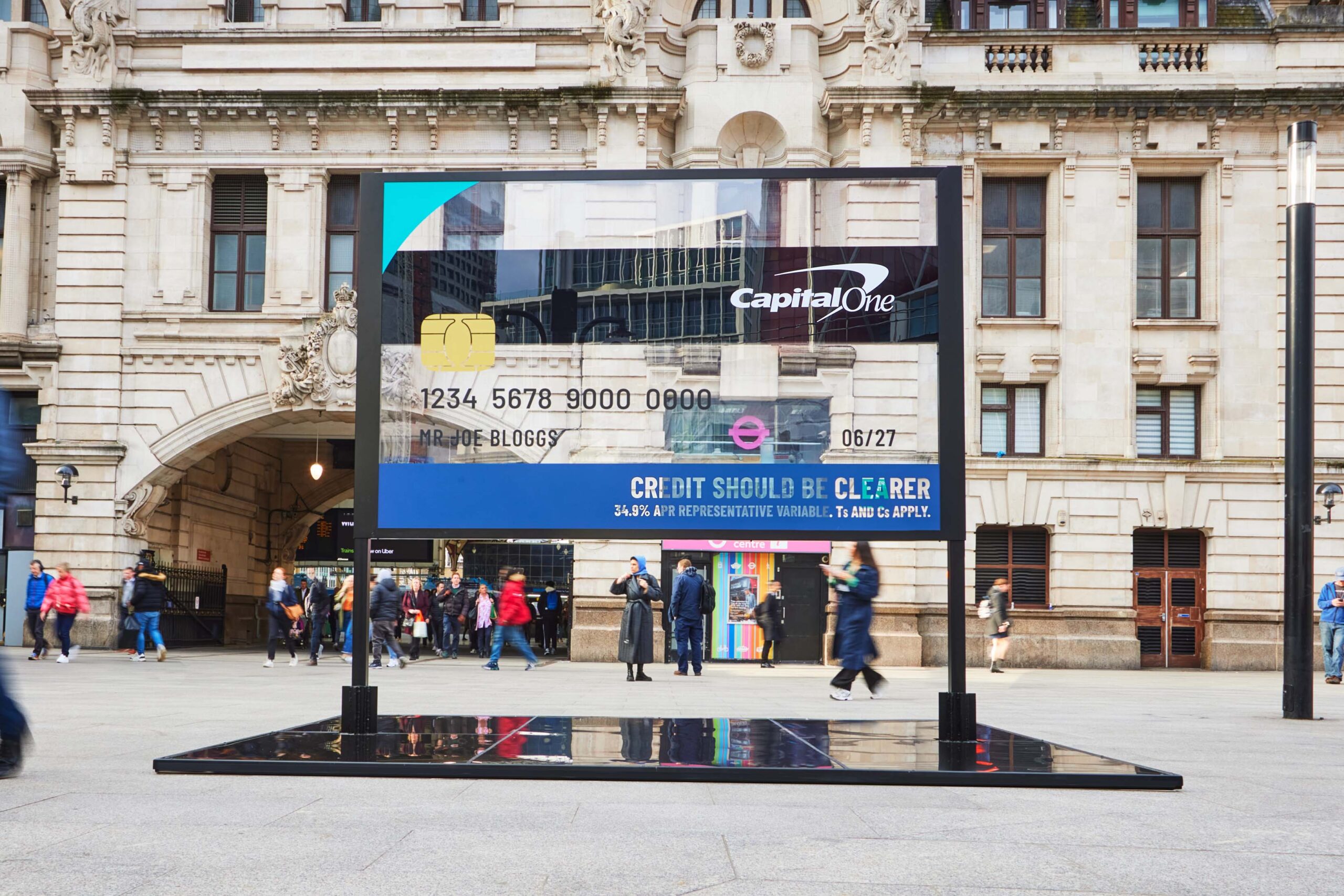 A transparent billboard installation at Victoria Station in London shaped like a large credit card, with people walking by, part of Capital One's 'Credit Made Clearer' campaign.