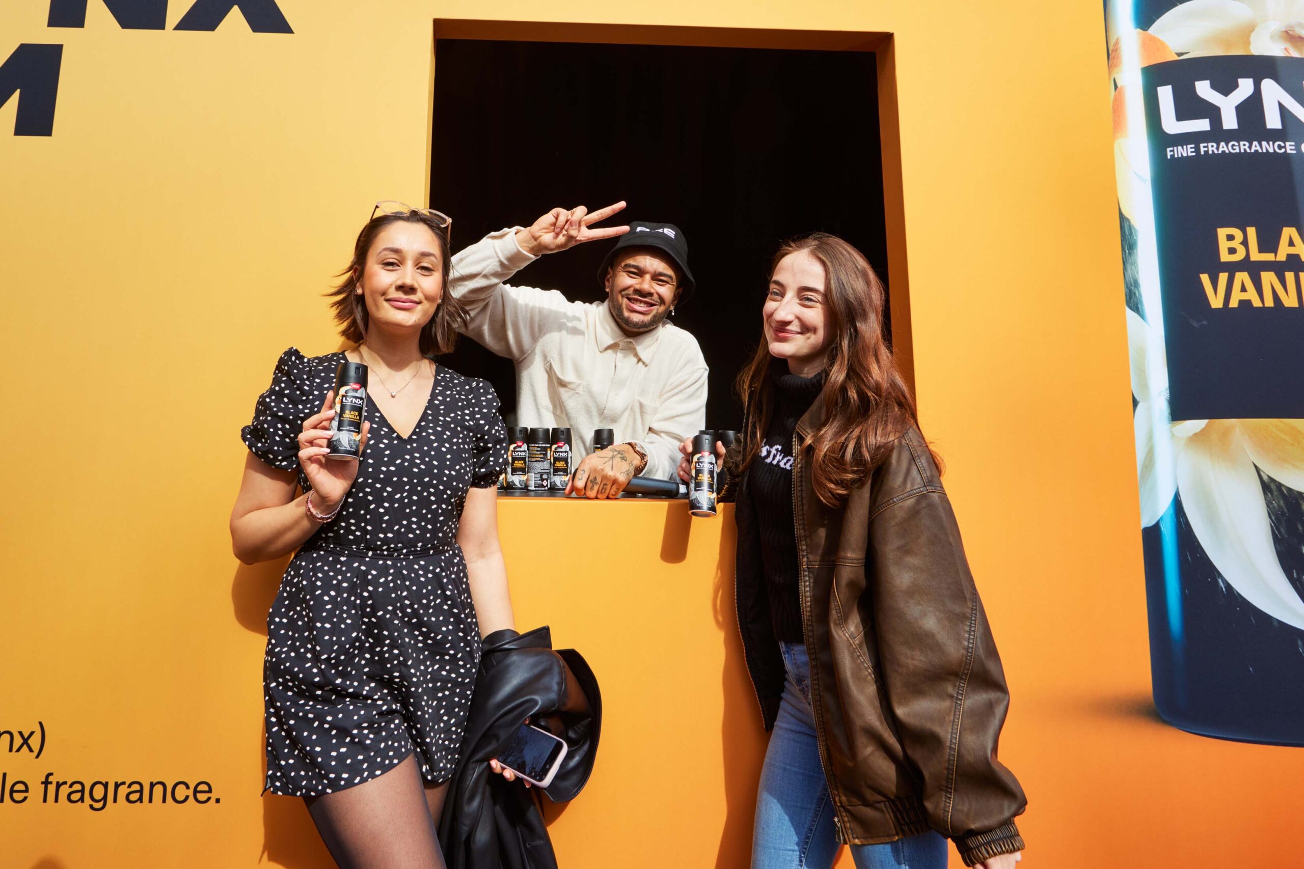 A yellow interactive billboard with a hatch. Wes Anderson is inside the hatch, handing out free samples to two excited young women who are posing for a photo with him.