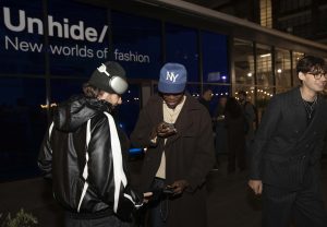 Three attendees at Lufthansa's "Unhide New Worlds of Fashion" event, one wearing headphones and a leather jacket, another in a brown coat and blue cap, engaged in a conversation. The background features a glass facade with the event's branding in vinyl and a warmly lit crowd indoors.